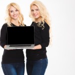Two beautiful smiling young blonde sisters twins showing blank laptop computer screen over white background